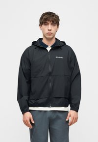 Νέος άνδρας που φορεί μαύρο φούτερ με φερμουάρ και κουκούλα της Columbia πάνω από λευκό πουκάμισο και γκρι παντελόνι, στέκεται μπροστά σε απλό φωτεινό φόντο.