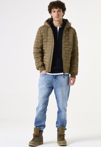 Chaqueta acolchada de color verde oliva con diseño acolchado, capucha, cremallera frontal, suéter de punto azul marino debajo, vaqueros azul claro y botas de senderismo marrones.