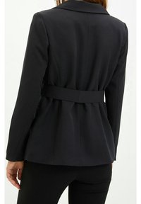 Musta räätälöity bleiseri, jossa istuva malli, leveät kaulukset ja vyötäröllä vyö. Kangas näyttää sileältä ja siinä on jämäkät saumat.