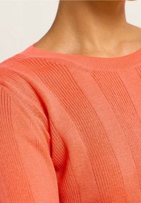 Close-up of neck and shoulder wearing a coral knit sweater with vertical ribbed patterns and a round neckline.