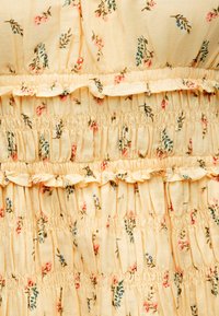Ανοιχτό κίτρινο ύφασμα με μικρά ροζ και μπλε λουλουδάτα σχέδια, που διαθέτει ελαστικό πιετάρισμα και οριζόντιες ραφές με βολάν.