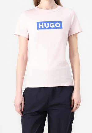 Person iført en lyserød T-shirt med korte ærmer med en blå rektangulær logo med teksten "HUGO" og mørkeblå bukser, stående mod en hvid baggrund.