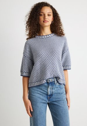 Young woman with curly hair wearing a blue and white textured knit sweater and high-waisted light blue jeans against a plain background.