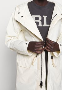 Krémově zbarvená, voděodolná parka se zapínáním na zip, nastavitelným stahováním v pase a velkými kapsami. Nositá přes tmavě šedé tričko s výrazným nápisem.