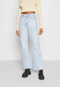 Jeans acampanados de un azul claro y cintura alta, con cinco bolsillos y cierre frontal con botón. Combinados con una blusa ceñida de color beige con textura acanalada.