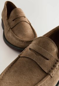 Mocasines de ante marrón con puntera redondeada, costuras decorativas y una banda elevada en la parte superior. Suela de goma negra. Textura suave.