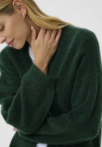 Ciemnozielony, puszysty sweter o luźnym kroju, z szerokimi rękawami i głębokim dekoltem w serek. Tekstura wygląda na miękką, z prążkowanymi mankietami. Brak wzorów i dodatków.