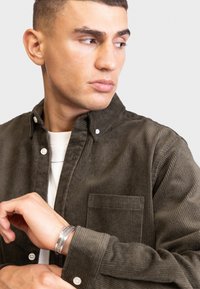 Olijfgroene corduroy overhemd met een knoopsluiting kraag, borstzak en subtiele textuur. Model draagt een wit shirt eronder. Zilveren armband zichtbaar.