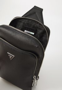 Crna sintetička torbica preko tijela s više zatvorenih pregrada i srebrnim hardverom. Unutra se nalazi obloga s ponavljajućim "GUESS" logotipom.