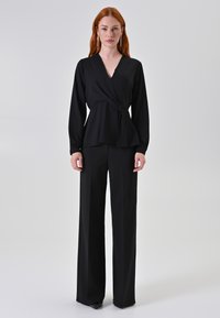 Blusa nera avvolgente con maniche lunghe, scollatura a V e vita arricciata, abbinata a pantaloni neri a vita alta e gamba larga. Tessuto morbido su tutta la superficie.