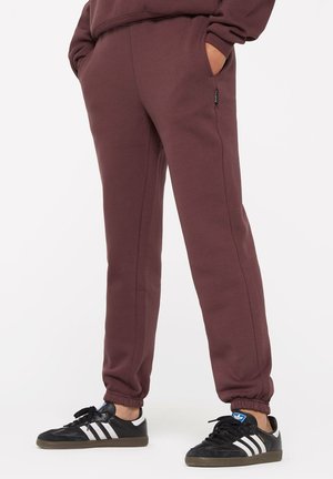 Bordeauxfarbene Sweatpants mit elastischen Bündchen, ausgestattet mit seitlichen Taschen und gesteppten Details. Dazu schwarze Sneakers mit weißen Streifen.