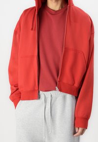 Roter, kurz geschnittener Hoodie mit einem Reißverschluss vorne, zwei Taschen und gerippten Manschetten, über einem roten Rundhalsausschnitt-T-Shirt und grauen Jogginghosen getragen.