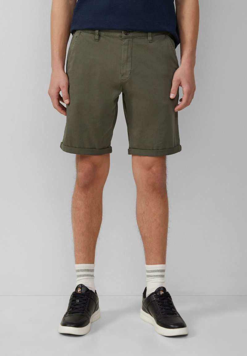 Shorts kaki avec revers, fabriqués en tissu léger. Associés à des baskets noires et des chaussettes blanches rayées, se tenant devant un fond neutre.