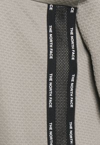Tessuto grigio texturizzato con una fascia in rete nera che presenta la scritta ripetuta in bianco "THE NORTH FACE", evidenziando il marchio e i dettagli di design.
