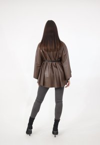 Giacca marrone in ecopelle con design plissettato, vita con cintura, maniche larghe e texture liscia, abbinata a pantaloni grigi aderenti e tacchi.