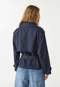 Marinblå kort trenchcoat med knappade axelpartier, midjebälte och slät textur. Bärs över ljusblå jeans.