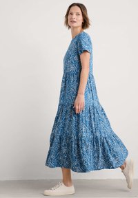 Robe maxi à motifs floraux bleus avec des manches courtes, design à volants, fabriquée en tissu léger. Portée avec des baskets blanches décontractées.