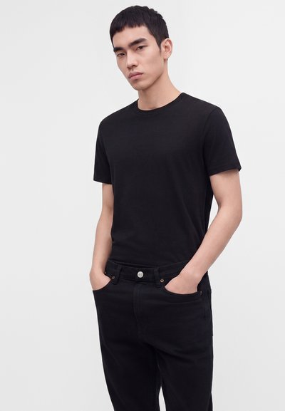 Jeune homme portant un t-shirt noir uni à manches courtes et un pantalon noir, les mains dans les poches, debout devant un fond clair uni.