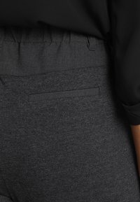 Pantaloni grigio scuro con una texture di tessuto liscia, una tasca posteriore piatta e una cintura elasticizzata, abbinati a una maglietta nera con maniche arrotolate.