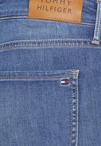 Jeans de denim con una etiqueta de cuero marrón claro con el nombre 'TOMMY HILFIGER' en relieve. Tejido azul claro con costuras doradas y pequeño logo en rojo, blanco y azul.