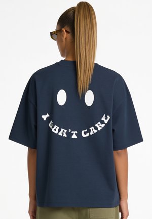 Woman with long blonde ponytail wears navy blue oversized t-shirt with white smiley face and text "I DON'T CARE" on the back.
