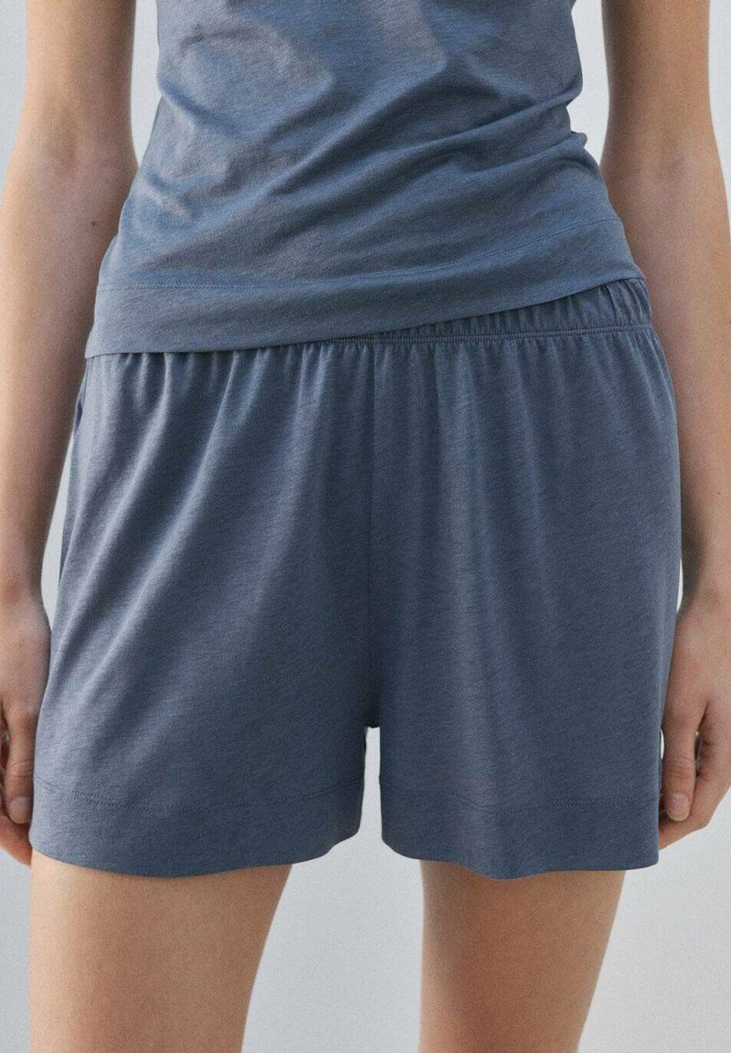 Femme portant un short à taille élastique foncé en tissu doux et ample, et un haut décontracté assorti, debout les mains détendues le long du corps.