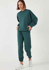 Felpa teal e pantaloni coordinati realizzati in tessuto morbido. La parte superiore presenta un collo rotondo e spalle scese, mentre i pantaloni hanno polsini elasticizzati.