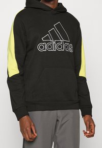 Felpa nera con cappuccio e dettagli gialli, caratterizzata da un grande logo Adidas bianco, tasca kangaroo frontale e polsini e orlo a coste.