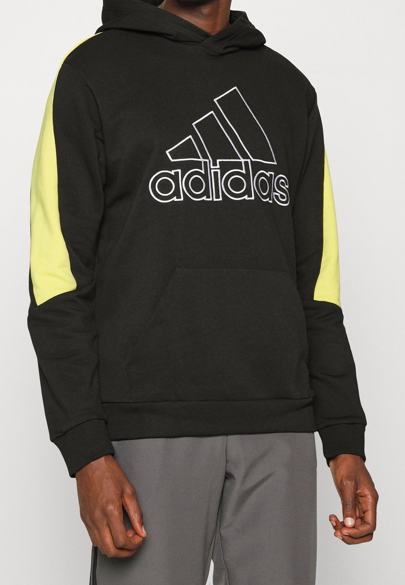 Felpa nera con cappuccio e dettagli gialli, caratterizzata da un grande logo Adidas bianco, tasca kangaroo frontale e polsini e orlo a coste.