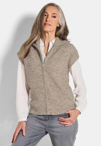 Beige gerippter Strickwesten mit Stehkragen und kurzen Ärmeln, mit Reißverschluss vorne, über einem weißen Hemd getragen, kombiniert mit grauen Jeans.