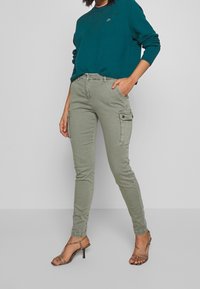 Lichtgroene cargobroek met een slanke pasvorm, meerdere zakken en knoopsluitingen, gecombineerd met een teal trui en sandalen met bandjes.