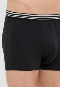 Melnas kokvilnas bikses bokseris ar svītrotu elastīgu jostu, cieši pieguļošas un ar šūšanas detaļām gar šuvēm.