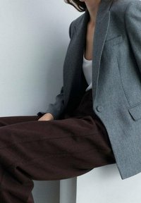 Harmaa räätälöity bleiseri lovikauluksilla, yhdistetty tummanruskeisiin leveälahkeisiin housuihin, joissa on sileät tekstuurit ja jäsennelty siluetti.