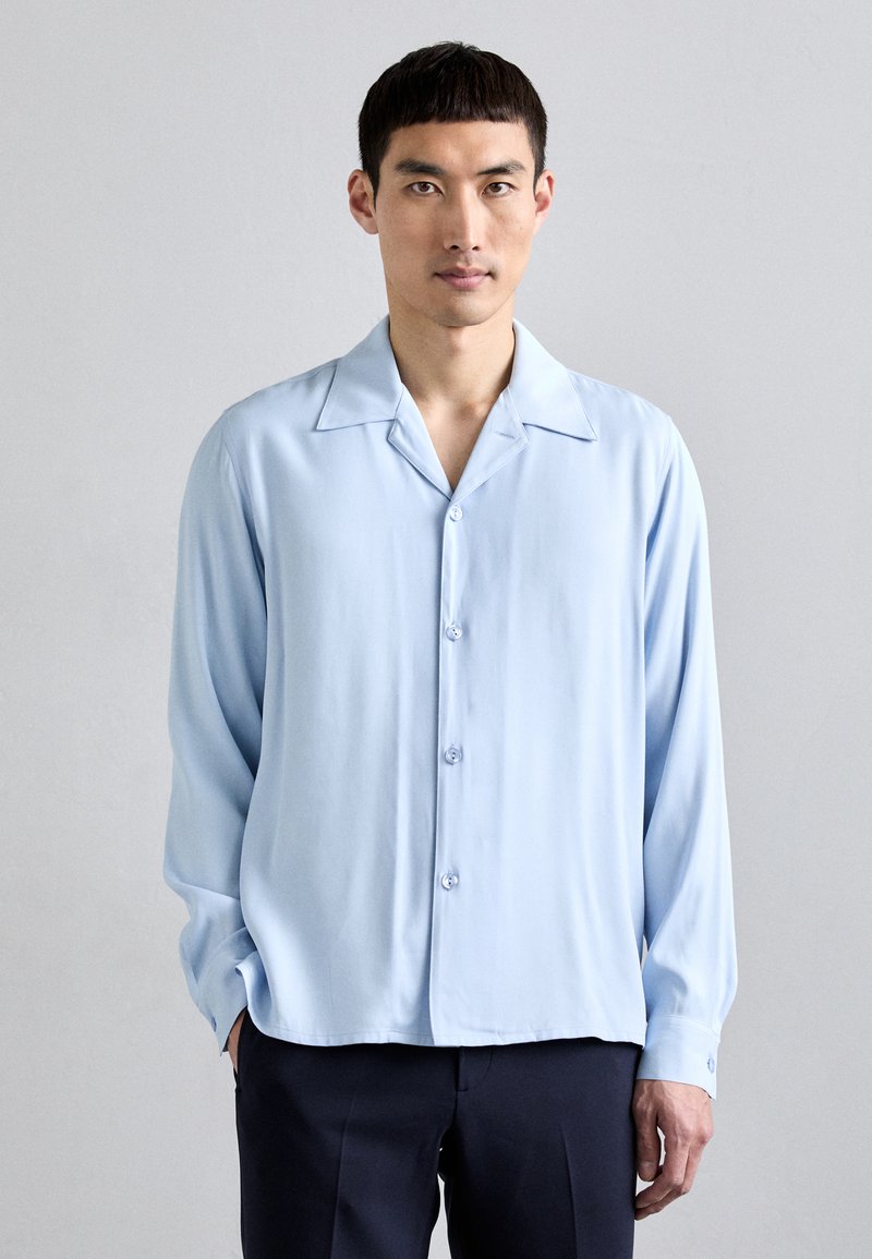 Lichtblauwe blouse met lange mouwen, met een notch-kraag, knoopsluiting aan de voorkant en een gladde stoffen textuur. Voorzien van twee knopen op elke manchette.
