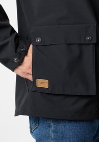 Zwarte jas met een pocket met twee drukknoopsluitingen, een bruine leren logo patch en een gladde, matte textuur.