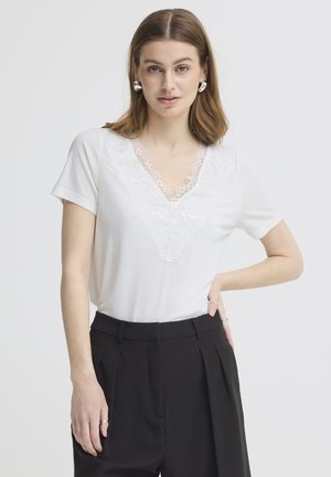Tânără cu păr castaniu până la umeri poartă un top alb de dantelă cu decolteu în V și pantaloni negri cu talie înaltă, pozând cu o mână pe șold pe fundal alb.