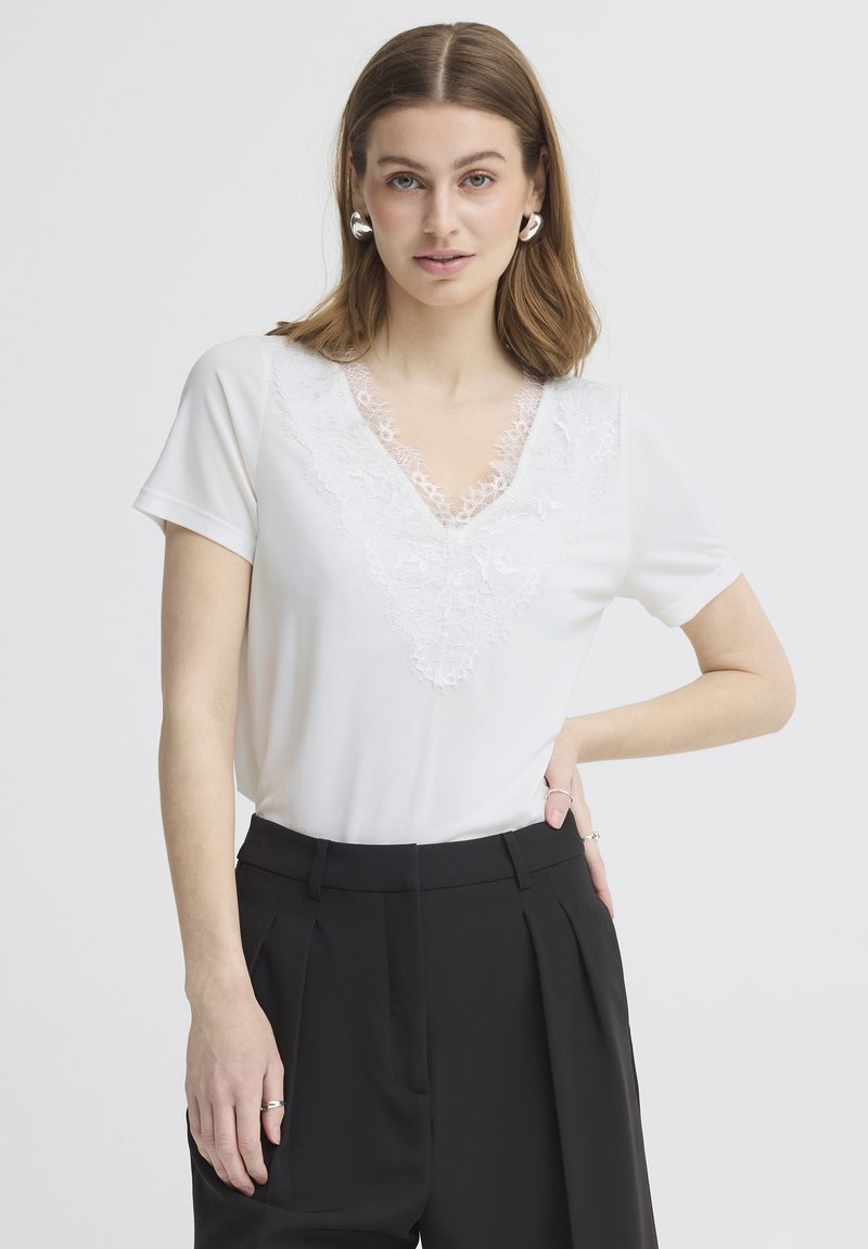 Jeune femme aux cheveux bruns mi-longs porte un haut en dentelle blanc à col en V et un pantalon noir taille haute, posant avec une main sur la hanche sur fond blanc.