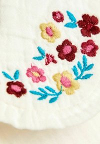 Flores bordadas coloridas en rojo, rosa y amarillo con hojas azules sobre una suave tela blanca, mostrando un acabado detallado y textura.