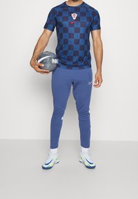 Marinblå kortärmad tröja med rutigt mönster, prydd med ett kroatiskt emblem och en röd Nike-logotyp. Kombinerad med blå joggers och Nike-skor.