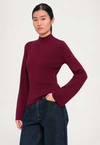 Sötét hajú nő, aki testhezálló bordó garbót és sötétkék farmert visel, az egyik kezét zsebében tartva pózol egy fehér háttér előtt.