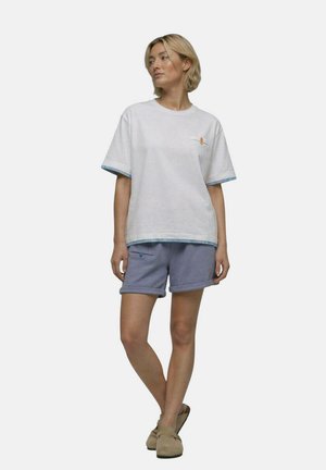 Jeune femme debout, jambes croisées, portant une chemise blanche à manches courtes, un short bleu et des chaussures beiges, regardant sur le côté sur un fond uni.