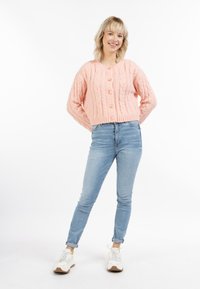 Rosa kabelstickad cardigan med sex knappar, kombinerad med ljusblå slim-fit jeans och vita sneakers med gula accenter.