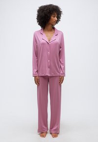 Γυναίκα όρθια, ξυπόλυτη, φορώντας φαρδιά, μωβ πουκάμισο με μακριά μανίκια και παντελόνα πιτζάμες, κοιτάζοντας πλάγια.