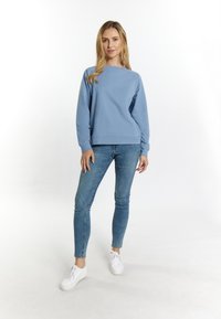 Világoskék pulóver, kerek nyakkivágással, bordázott mandzsetta és szegély, kék skinny farmerral és fehér sportcipővel viselve, sima háttér előtt.