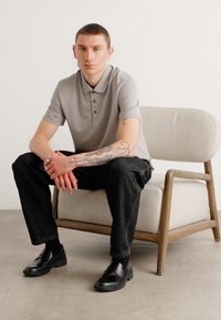 Grå polo skjorte, svarte jeans og svarte sko. Sitter på en trestol med tekstilbehandling; casual, avslappet holdning.