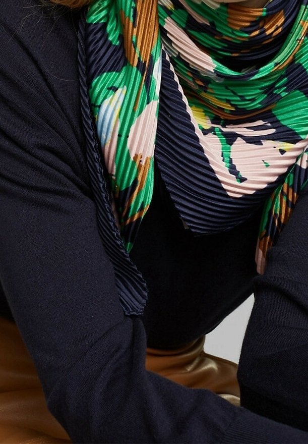 Personne portant un pull bleu marine foncé et une écharpe plissée avec des motifs floraux abstraits verts, bleu marine, beige et marron.