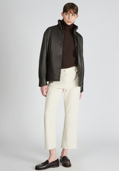 Giacca di pelle nera, maglione a collo alto marrone, pantaloni cropped crema e mocassini scuri; texture morbide e colori a contrasto.