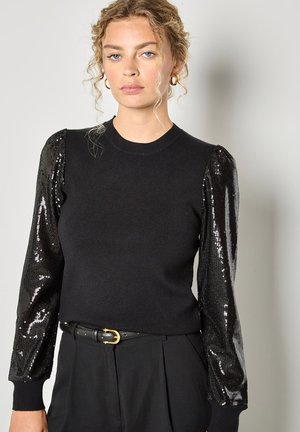 Zwarte trui met pailletten opgeblazen mouwen en een ronde halslijn, gecombineerd met hooggesneden broek en een zwarte riem met gouden gesp.