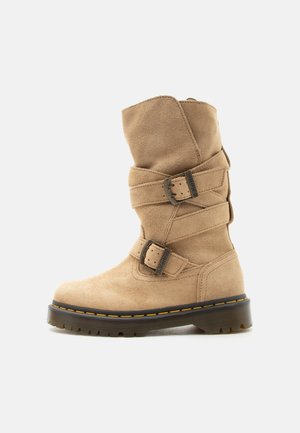 Botte mi-mollet en daim couleur tan avec deux sangles ajustables et boucles métalliques, avec une semelle noire épaisse et surpiqûres jaunes.