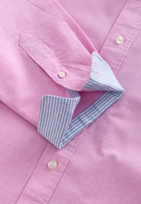 Chemise rose à boutons avec une manchette repliée, dévoilant une doublure intérieure rayée bleue et blanche ainsi que des boutons blancs sur le tissu.